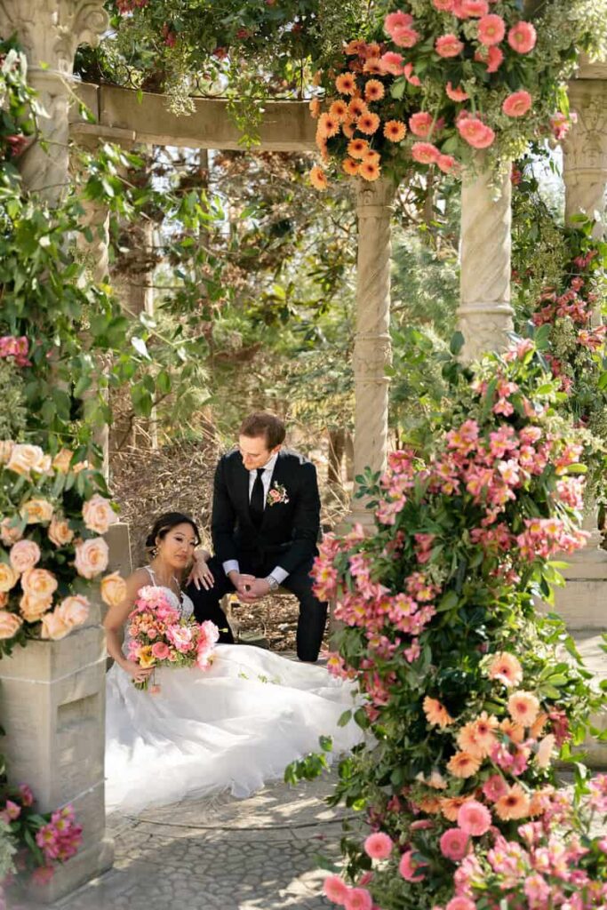 Bride and groom surrounded by incredible fresh florals, bride is holding a lush bridal bouquet of fresh tulips and seasonal florals by Intrigue Designs at Crystal Plaza wedding hosted by Styled Shoots Across America.