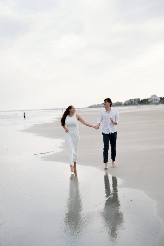 Engaged couple running barefoot through the surf during a beach engagement session in South Carolina.

