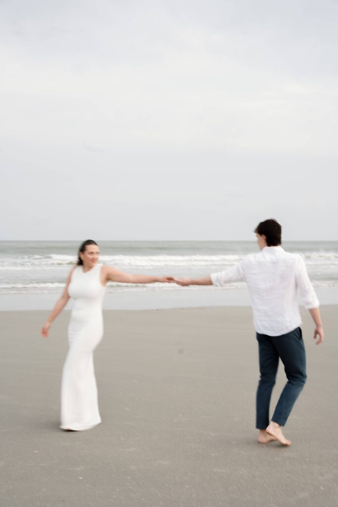 Couple holding hands and walking along Folly Beach during their Charleston engagement session